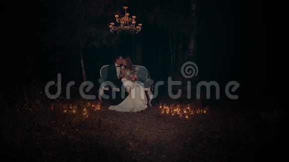 新郎和新娘在晚上拥抱和亲吻坐在黑暗森林的沙发上视频的预览图