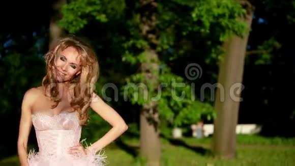 年轻的金发女孩穿着粉红色优雅的裙子站在树林里视频的预览图
