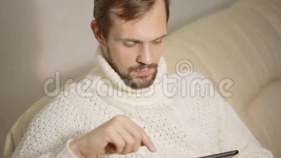 一个长胡子的年轻人穿着夹克坐在沙发上拿着平板电脑视频的预览图