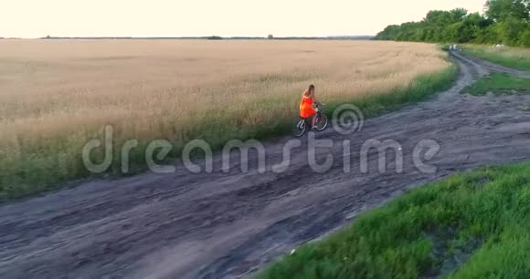 穿裙子的女孩骑着自行车沿着麦田向无人机射击从高处看美丽的风景视频的预览图