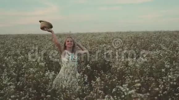 手里拿着帽子的女人走在一片开着野花的田野里视频的预览图