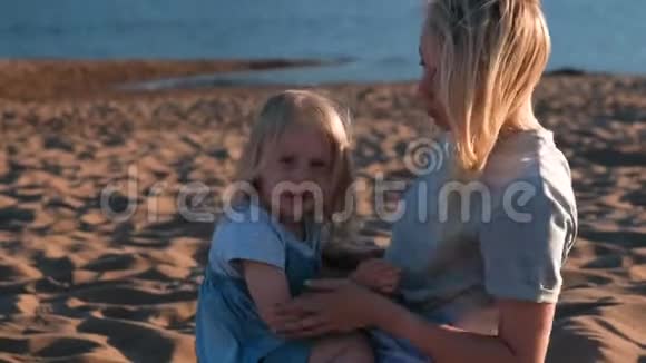 美丽的金发妈妈和女儿在日落时在海滩上拥抱视频的预览图