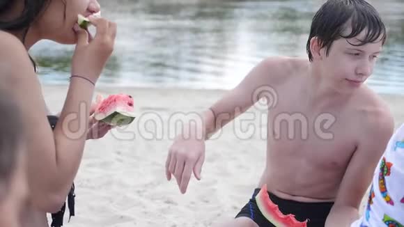 孩子们青少年在海滩上休息笑着吃西瓜慢动作视频男孩和女孩的生活方式视频的预览图