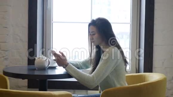 年轻的黑发女孩坐在靠近窗户的桌子上看着平板电脑视频的预览图