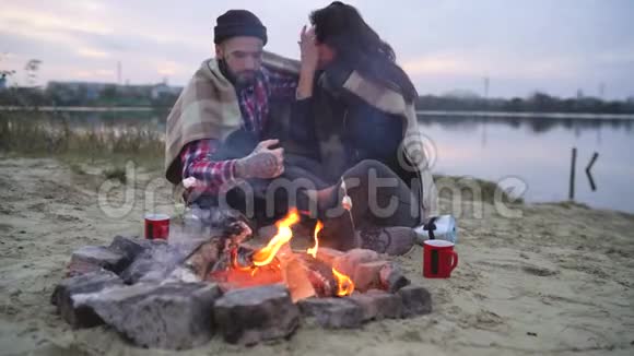 一对恋爱中的情侣坐在湖边的篝火旁聊着天喝着热茶视频的预览图