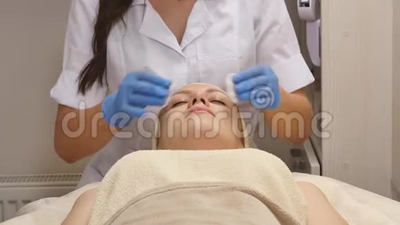 一个闭着眼睛的漂亮女人躺在美容师的桌子上皮肤科医生用棉花海绵做视频的预览图