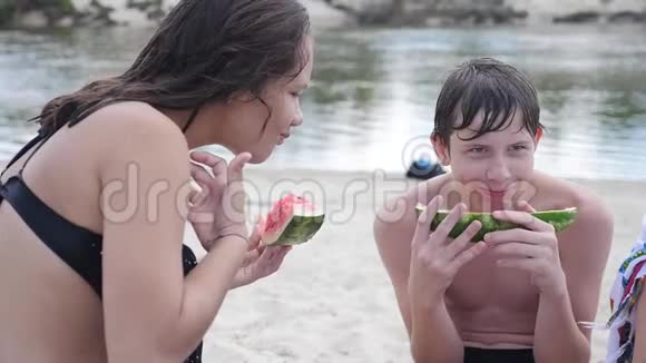 孩子们青少年在海滩上休息笑着吃西瓜慢动作视频男孩的生活方式和女孩视频的预览图