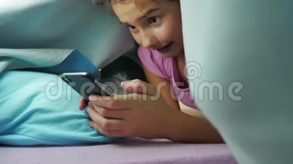 十几岁的女孩和夜狗在毯子下玩智能手机睡前的小女孩躺在床上冲浪视频的预览图