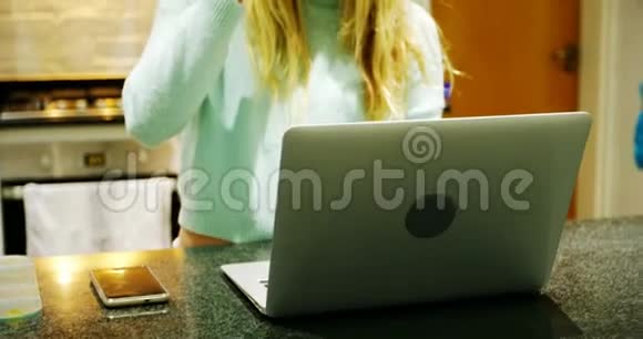 女人一边喝咖啡一边用厨房里的笔记本电脑视频的预览图