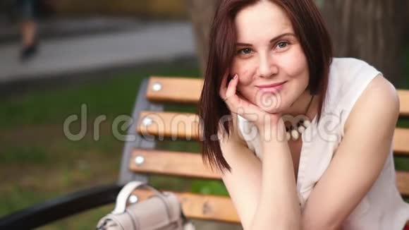 坐在夏日公园的长椅上美丽的年轻女子神秘地微笑着户外妇女的肖像视频的预览图