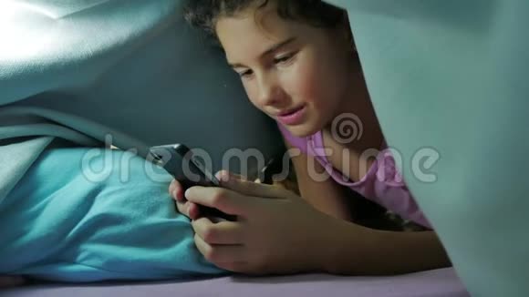少女之夜和狗在毯子下玩智能手机睡前的小女孩躺在床上冲浪视频的预览图