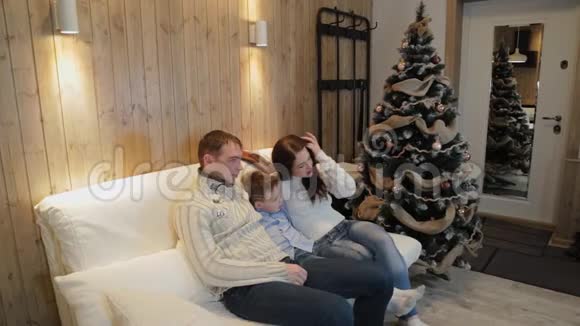美丽幸福的三口之家在沙发上微笑着在圣诞夜的圣诞树旁交谈视频的预览图