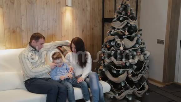 美丽幸福的三口之家在沙发上微笑着在圣诞夜的圣诞树旁交谈视频的预览图
