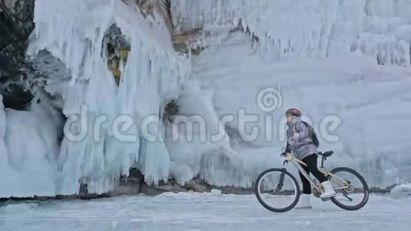 女人正在冰窟附近骑自行车有冰洞和冰柱的岩石非常美丽女孩穿着视频的预览图