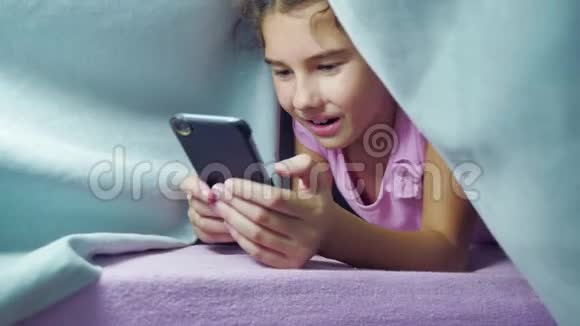 女孩少年和狗在夜里在智能手机里玩毯子睡前的小女孩躺在床上冲浪视频的预览图