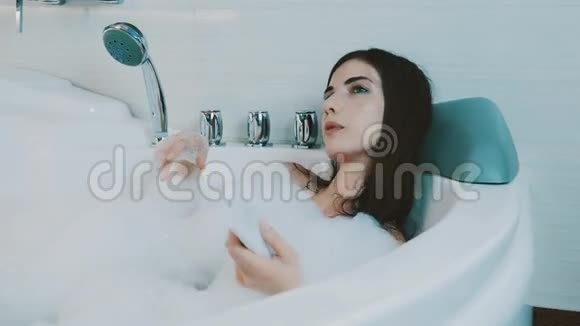布鲁内特女孩在浴室里洗了满是泡沫的澡通过控制器切换音乐视频的预览图