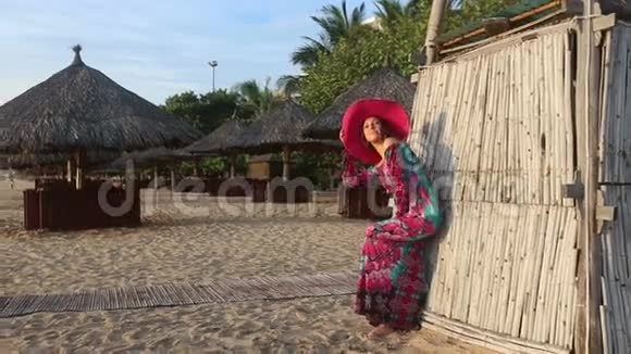 穿着长裙戴着红帽子的女孩在海滩竹塔边摆姿势视频的预览图