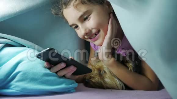 少女和狗在毯子下玩智能手机小女孩睡前躺在床上在室内冲浪视频的预览图