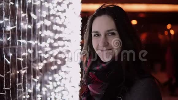 圣诞夜飘雪中年轻迷人的女人看着背景的灯光视频的预览图
