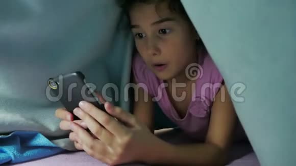 少女和狗在毯子下玩智能手机的夜晚睡前的小女孩躺在床上冲浪视频的预览图