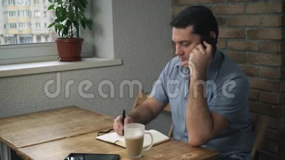 穿着蓝色衬衫的经理坐在咖啡馆里打电话在笔记本上记笔记桌上有一杯拿铁视频的预览图