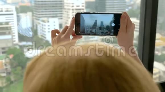 优雅的金发女人在电话里拍照女人从摩天大楼的窗户上拍摄风景视频的预览图