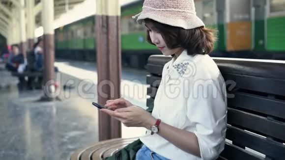 快乐的年轻女性坐在长凳上用智能手机在火车站聊天阅读和发短信视频的预览图