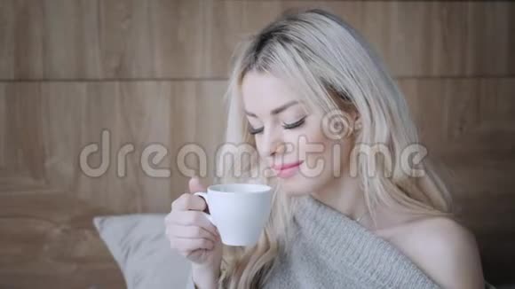 美丽的金发女人拿着陶瓷杯的茶或咖啡享受在床上喝茶看着窗户喝茶视频的预览图