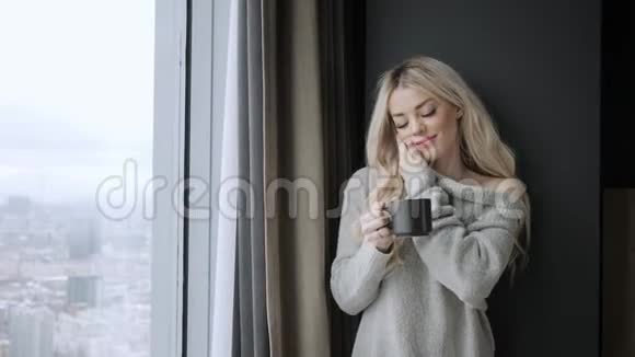 美丽的金发女人拿着陶瓷杯的茶或咖啡享受在床上喝茶看着窗户喝茶视频的预览图