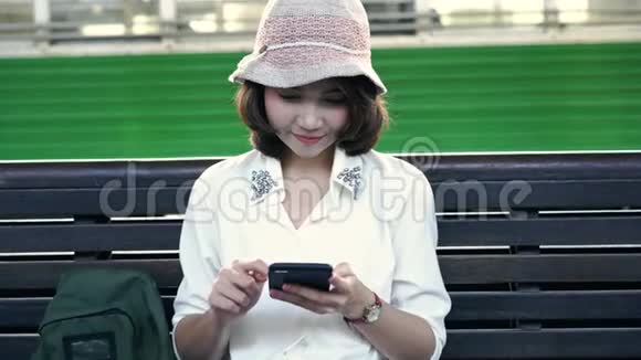 快乐的年轻女性坐在长凳上用智能手机在火车站聊天阅读和发短信视频的预览图