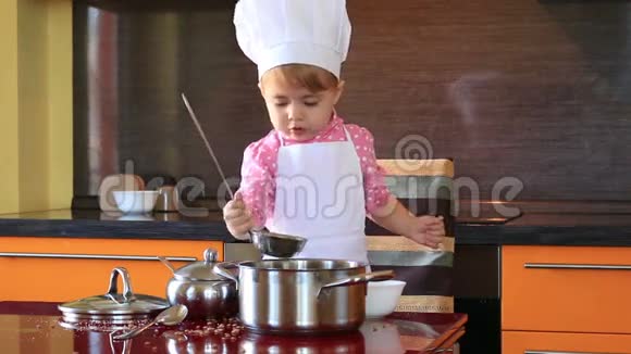 穿着厨师服的小可爱女孩在厨房里用勺子和平底锅玩视频的预览图