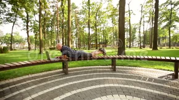 一个拿着笔记本电脑的商人躺在公园的长凳上视频的预览图