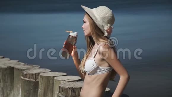 一个年轻的女人戴着夏帽穿着白色比基尼泳衣洗太阳浴喝瓶子里的水视频的预览图