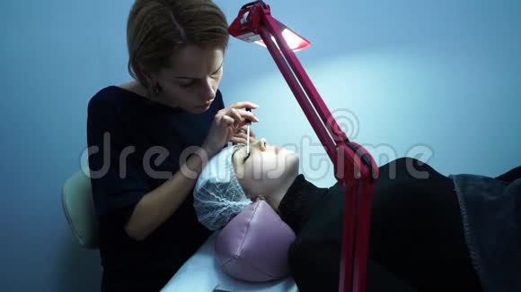 睫毛延伸化妆大师把睫毛伸向一个年轻女孩那个女孩躺着旁边有一盏灯视频的预览图