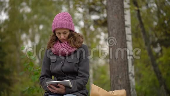 在秋天公园坐在长凳上时年轻的女人用平板电脑坐在长椅上的学生视频的预览图