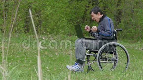 残疾男子惊喜成功胜利轮椅与笔记本电脑在轮椅上工作的自然绿色背景视频的预览图