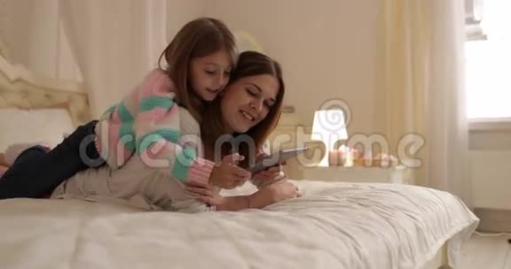 母亲带着女儿躺在床上用平板电脑快乐地微笑着年轻的家庭视频的预览图