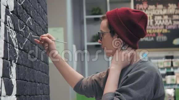 年轻的女画家在咖啡店的黑砖墙上做白色设计视频的预览图