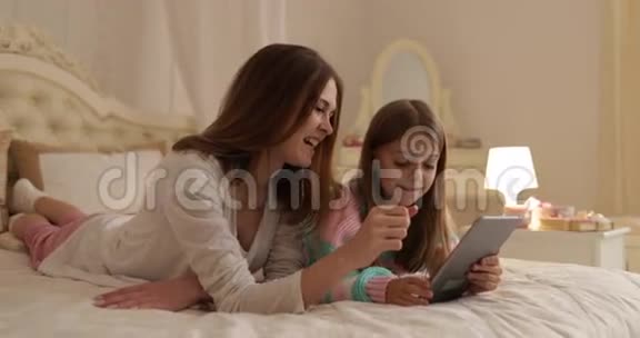 母亲带着女儿躺在床上用平板电脑快乐地微笑着年轻的家庭视频的预览图