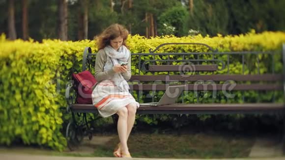 在美丽的绿色公园的长凳上使用智能手机放松的女人校园中的年轻千禧妇女做手势视频的预览图