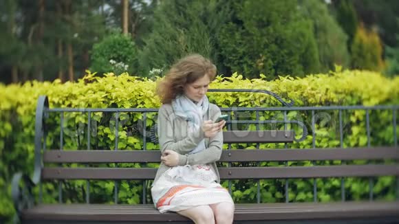 在美丽的绿色公园的长凳上使用智能手机放松的女人校园中的年轻千禧妇女做手势视频的预览图