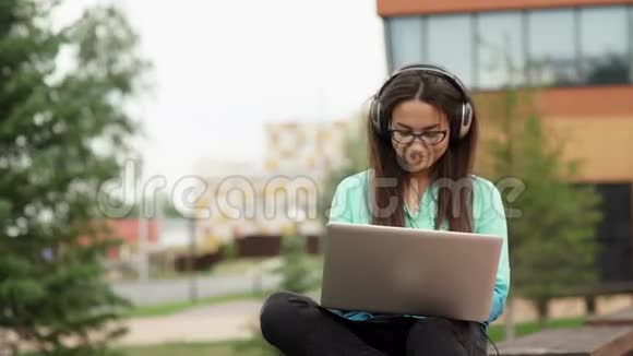 可爱的年轻女孩带着耳机坐在空地上带着笔记本电脑视频的预览图