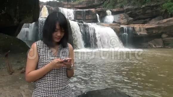 年轻女性使用智能手机户外活动坐在瀑布附近微笑视频的预览图