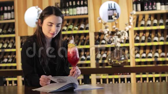 一个年轻女孩在一家时尚餐厅的桌子上喝一杯鸡尾酒一个年轻女孩在一张桌子上喝一杯红色异国情调的鸡尾酒视频的预览图
