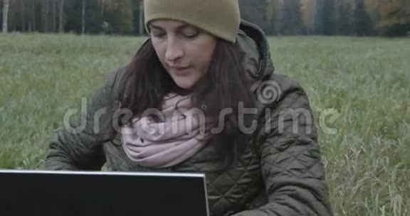 年轻的深色头发的女学生在大自然中做笔记本电脑美丽的秋日和女人在使用笔记本电脑时享受它视频的预览图