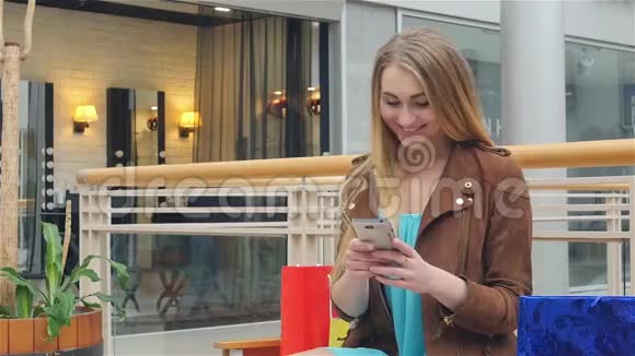 金发女人坐在购物中心带着电话视频的预览图
