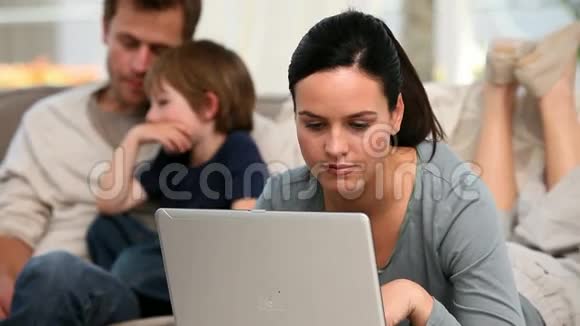 美丽的女人在她的电脑上而她的家人在沙发上视频的预览图