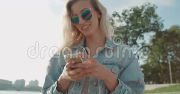 阳光明媚的一天年轻的金发女人戴着太阳镜在城市里用手机视频的预览图