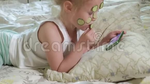一个漂亮的小女孩兴高采烈地在家里制作黄瓜面具并使用智能手机视频的预览图