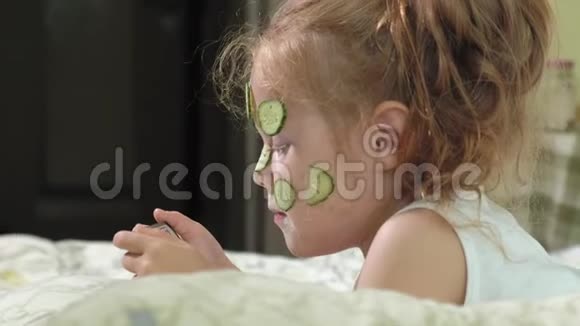 一个漂亮的小女孩兴高采烈地在家里制作黄瓜面具并使用智能手机视频的预览图